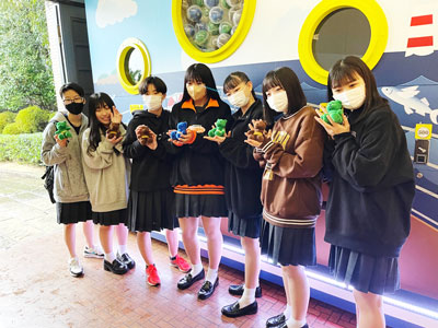 ぬいぐるみの景品を手にした制服姿のグループが、ガチャマシンの前で笑顔